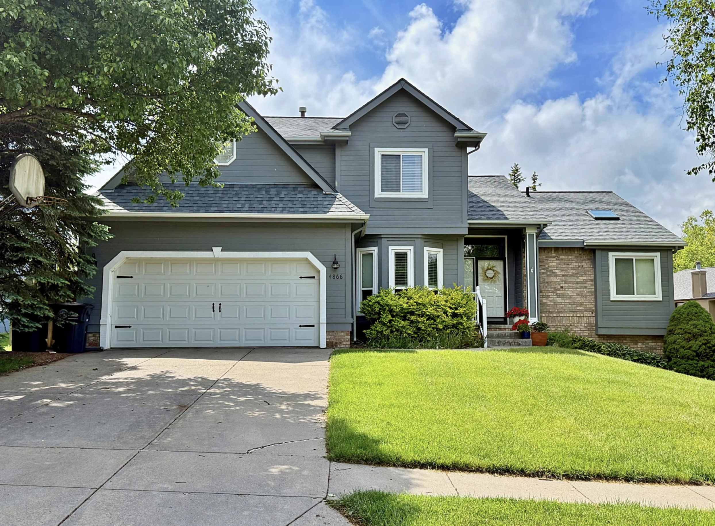 A two-story house painted in gray with white trim, a white garage door, green bushes and a lawn in front, surrounded by trees, under a partly cloudy sky.
