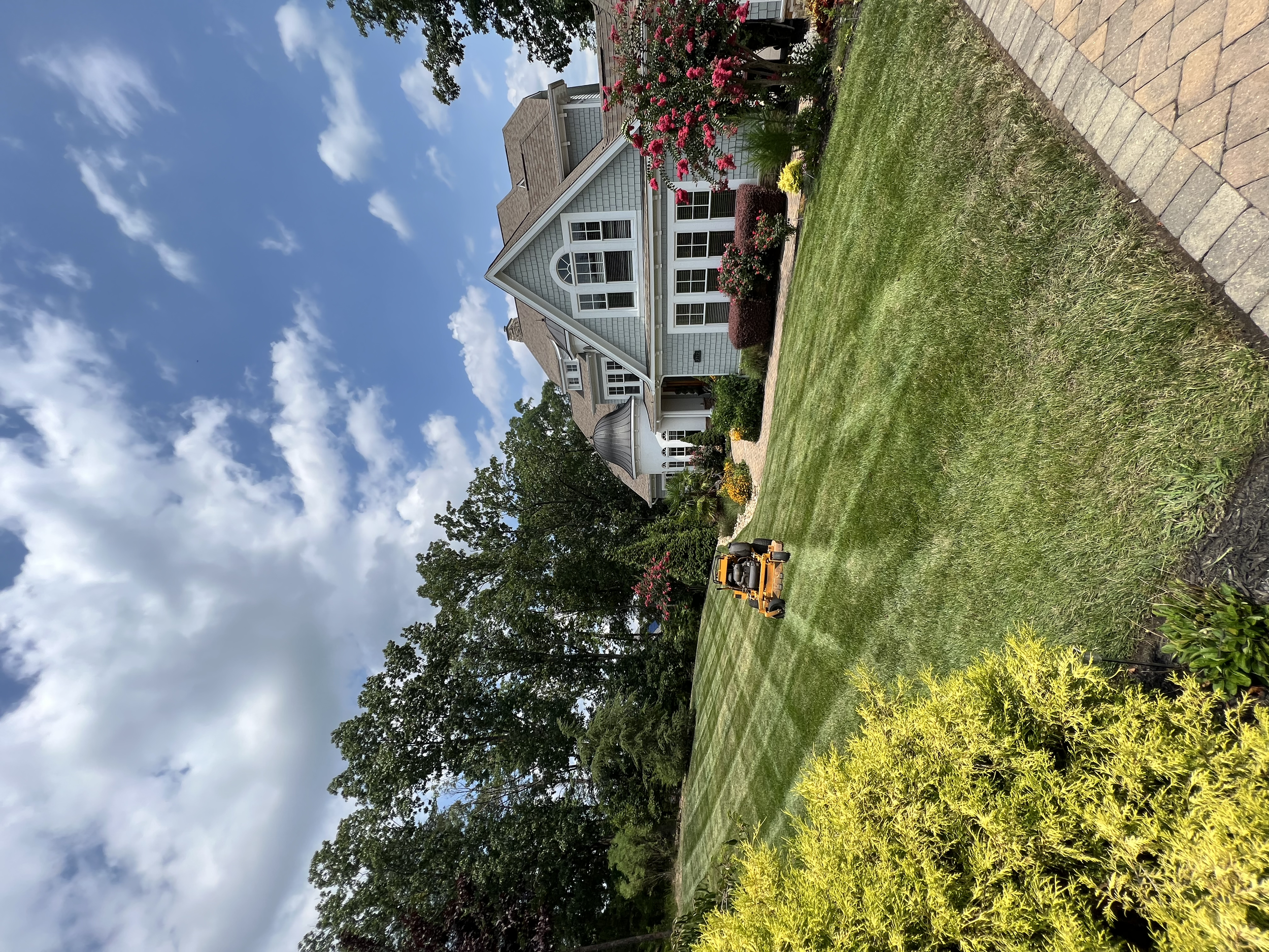 A manicured lawn being mowed with a yellow riding lawn mower outside a large, white, Victorian-style house with a porch, multiple windows, and a gable roof. The house is surrounded by trees and colorful flower beds under a partly cloudy sky.
