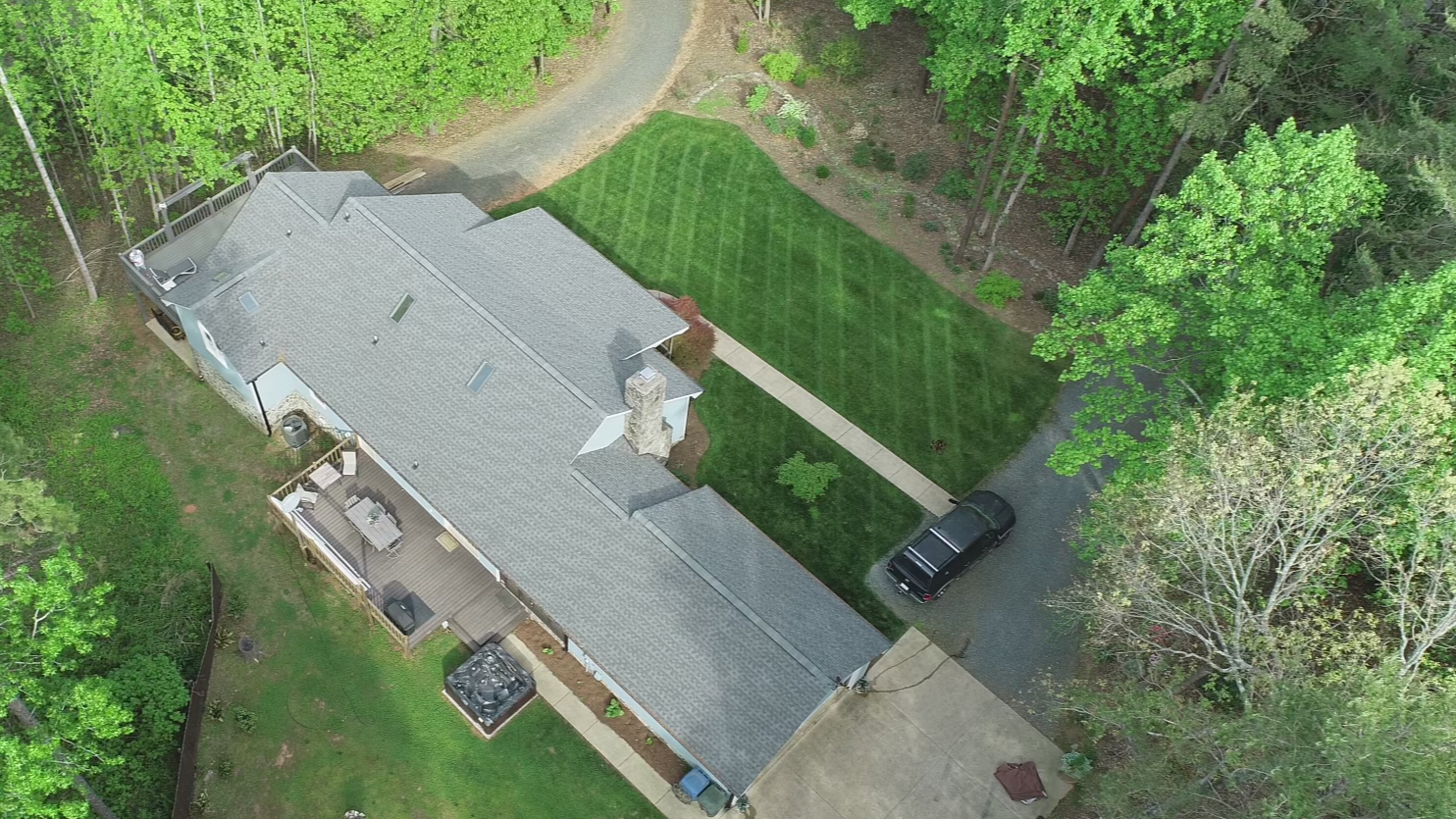 Aerial view of a suburban house with a gray roof, surrounded by lush green trees and lawns. The backyard features a deck with outdoor furniture, a fire pit, and a grill. A paved driveway with a black car leads to the house, and there is a well-mainta
