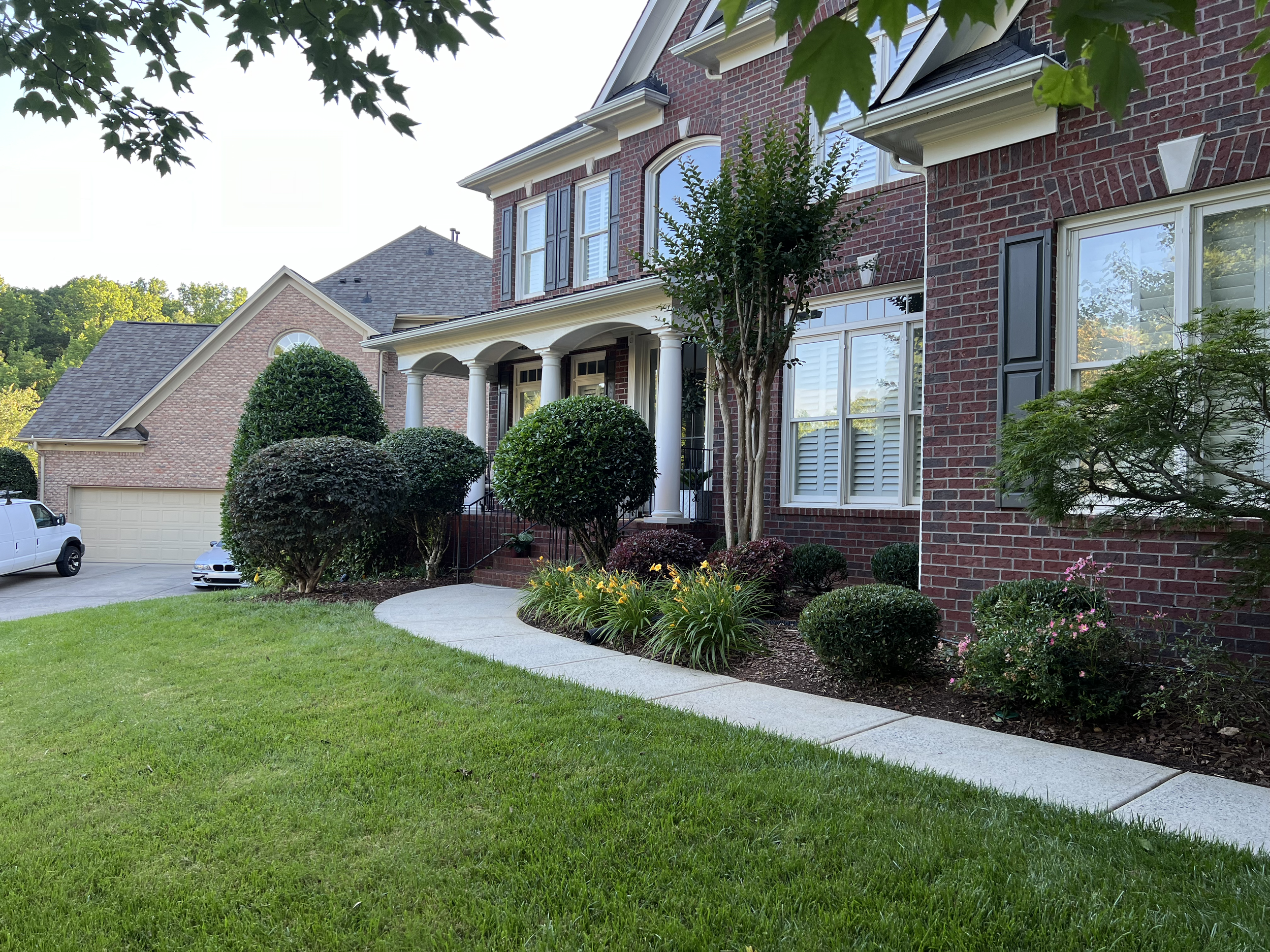 Front yard of a brick house with a curved concrete walkway, well-maintained green lawn, and landscaped bushes and flowers.
