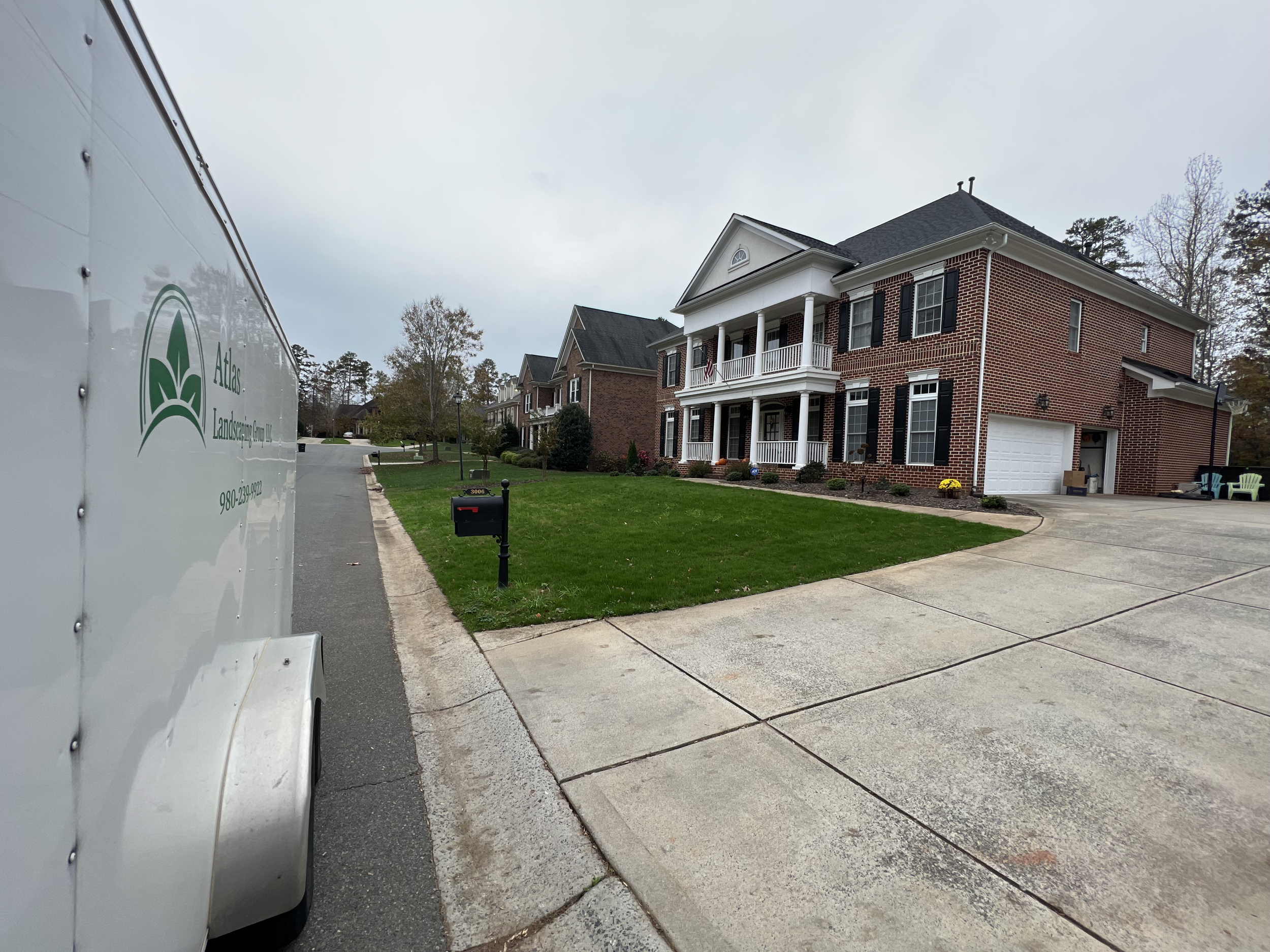 A front view of a large brick house with white columns and a porch, and a well-maintained lawn. A driveway and a sidewalk are visible, with outdoor chairs and a mailbox in the yard. Part of a white truck with a logo and text is on the left side of th