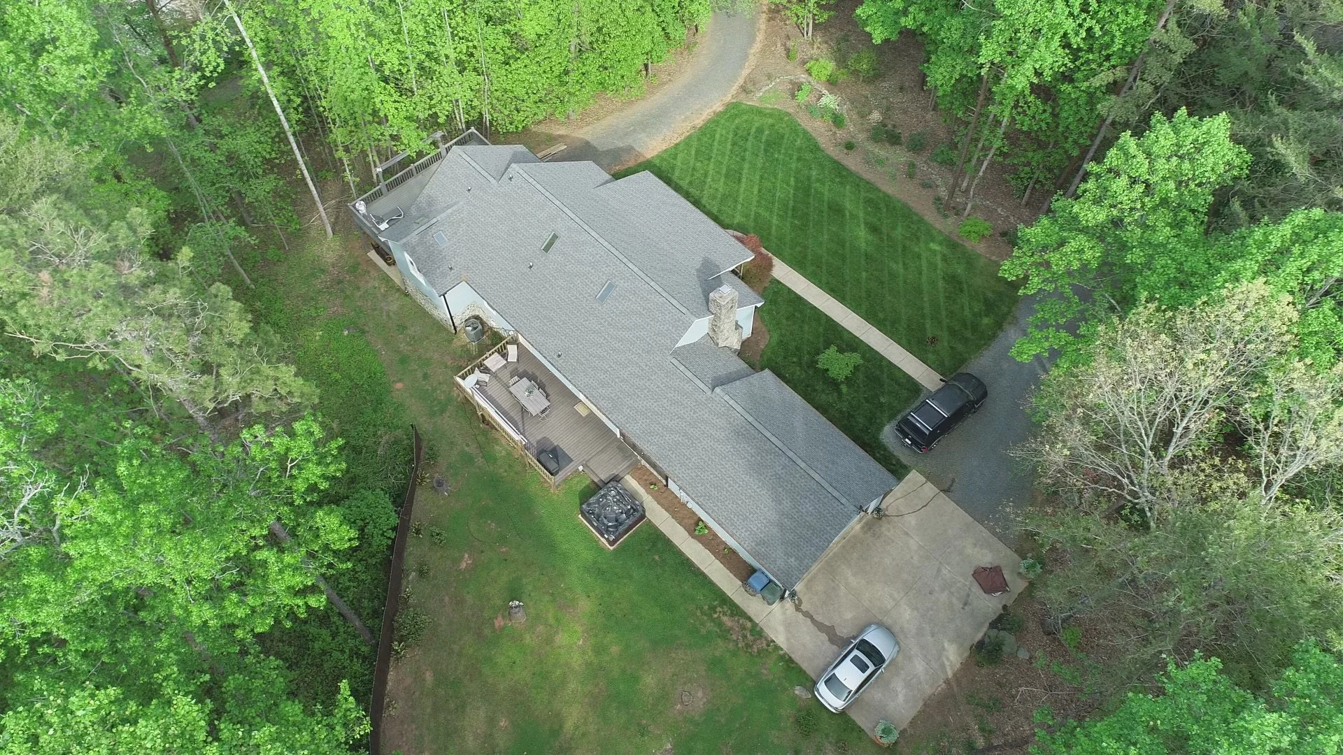 Aerial view of a house surrounded by green trees and a well-maintained lawn. The house has a grey roof, a backyard deck with outdoor furniture, and a driveway with three cars parked.
