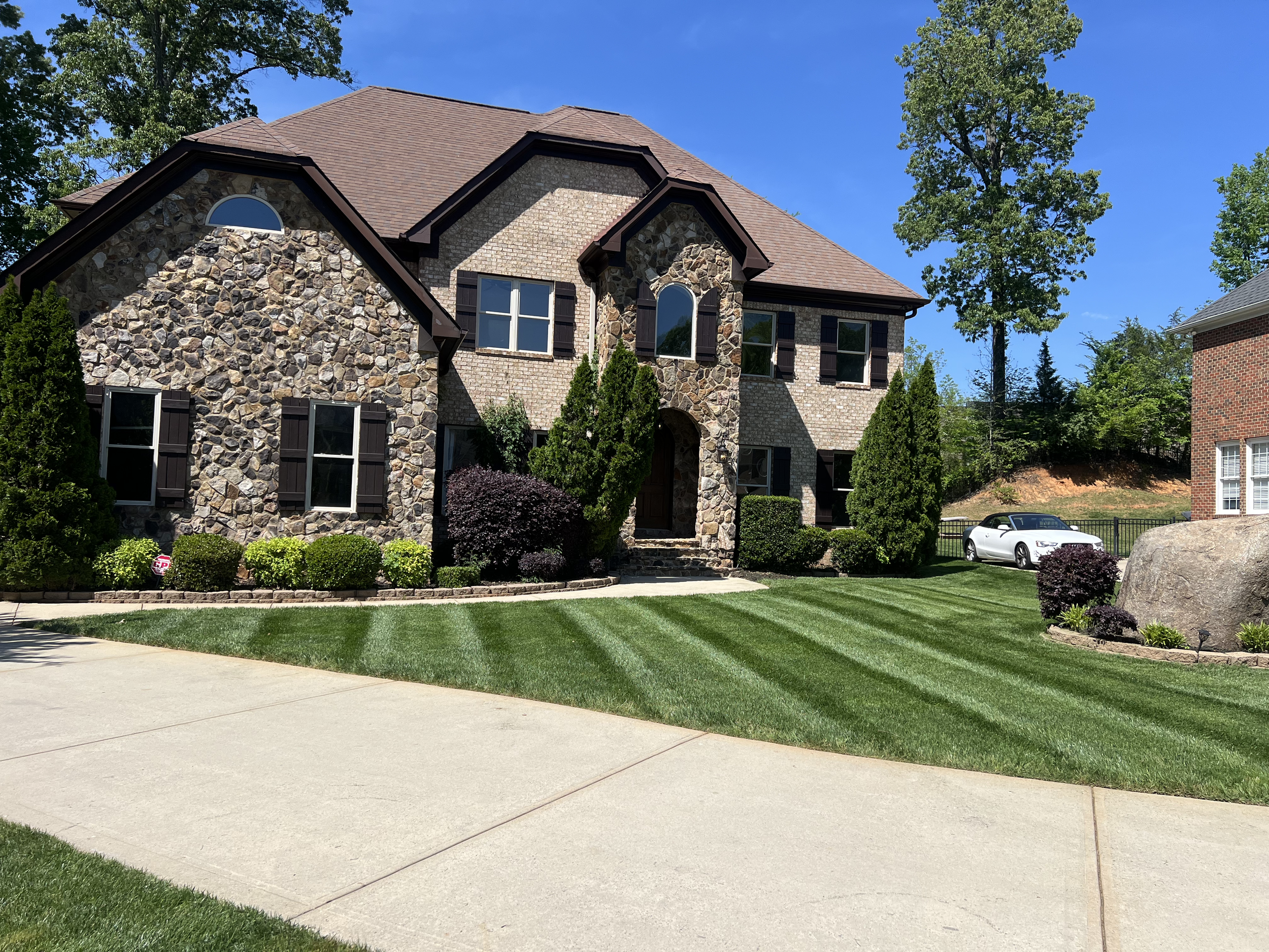 A two-story house with a stone and brick exterior, surrounded by a well-maintained lawn and trees, with a white car parked in the driveway.