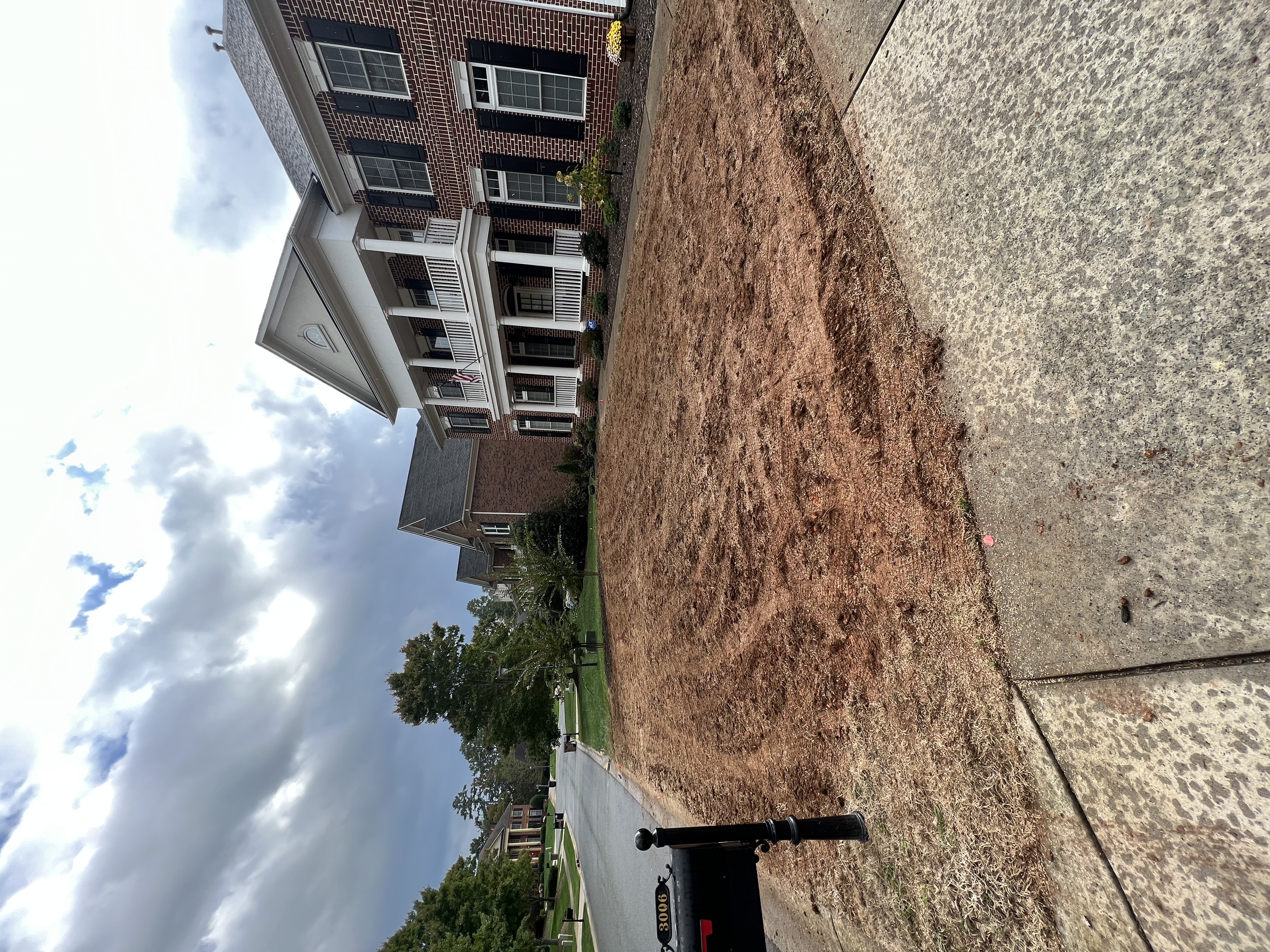 A residential street with brick houses, a sidewalk, and a small patch of dirt in the foreground. The sky is cloudy with patches of blue. There are trees and a lawn in the background.
