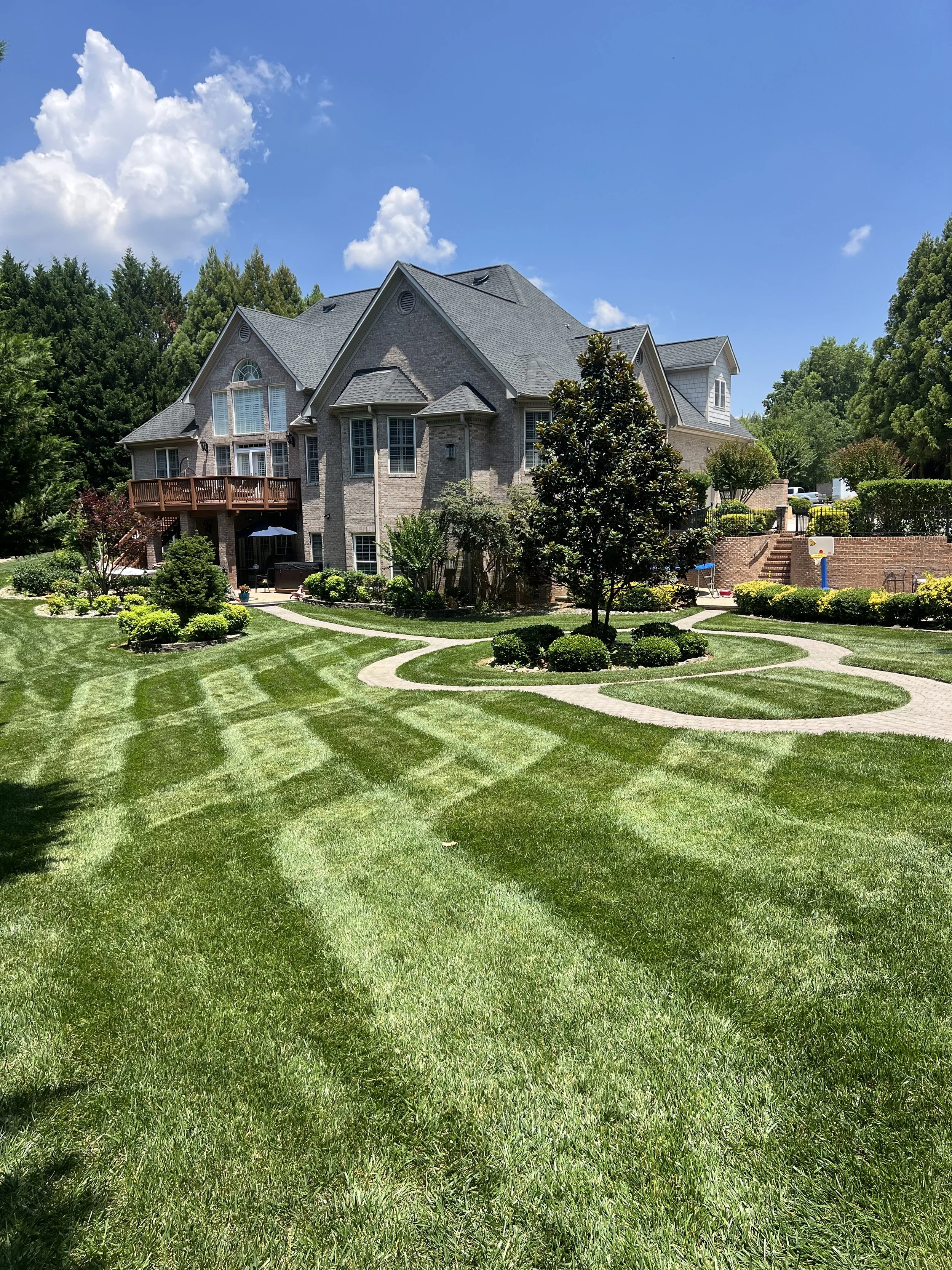 A large, well-maintained lawn with striped grass, leading up to a large brick house with multiple gables and windows. There are trees, bushes, and flowerbeds surrounding the house, with a clear blue sky overhead.