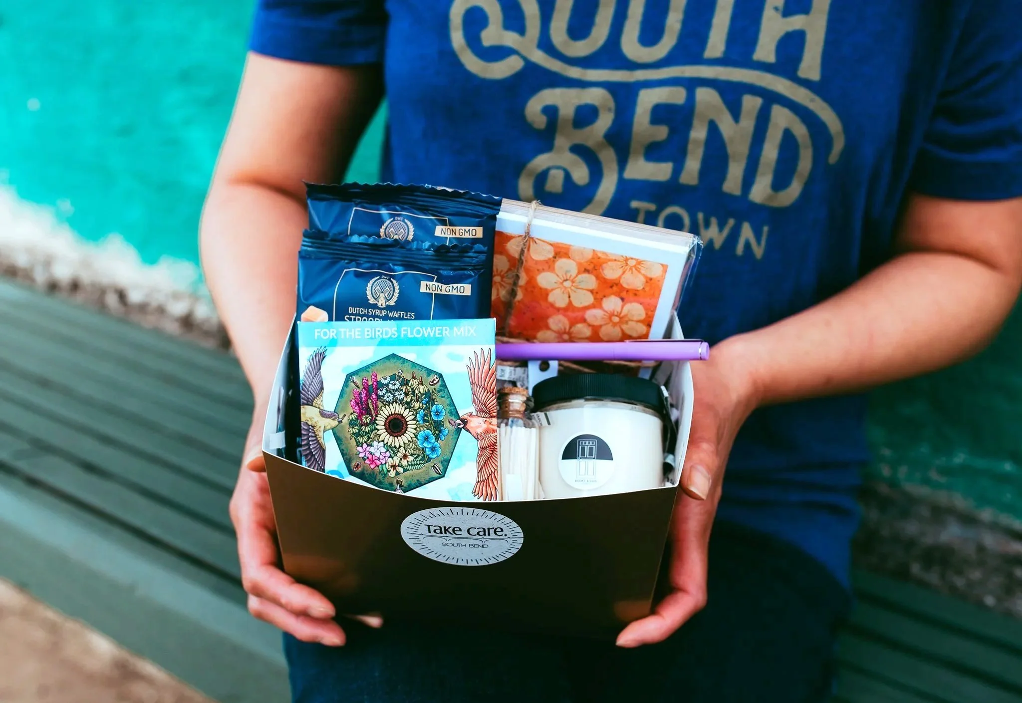 A person holding a black takeaway box with various items including seed packets, a candle, and a small notebook. The person is wearing a blue shirt with the word 'COUNTY' visible.