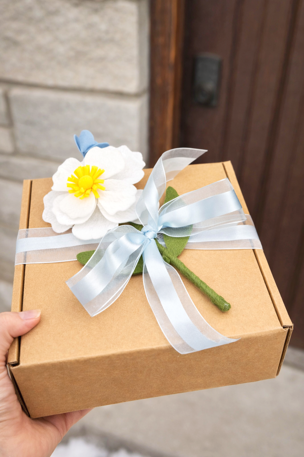 A hand holding a gift box decorated with a fabric flower, green leaves, and a white ribbon bow.