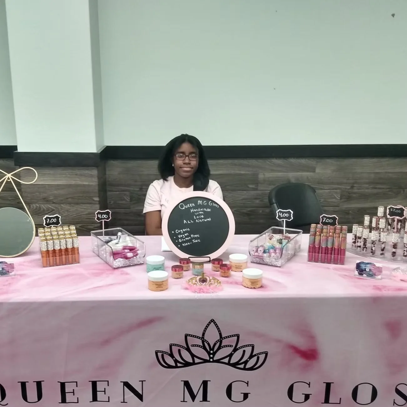 A young girl is sitting at a table with handmade cosmetic products and signs with prices, behind a pink tent with the label 'Queen MG Glos'.