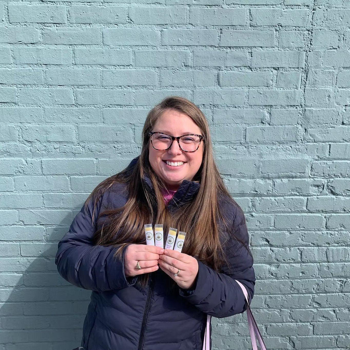 A smiling woman with long brown hair, glasses, and a dark jacket, standing against a light blue brick wall, holding four small bottles.