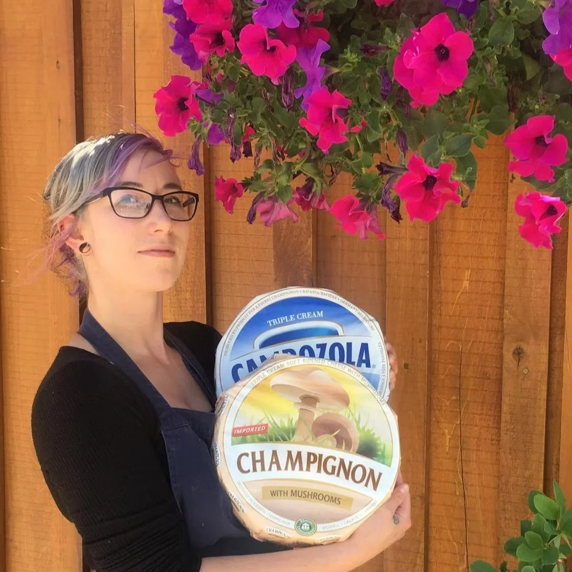 A woman with glasses and multi-colored hair holds two containers of cheese, one labeled 'Champignon with Mushrooms' and the other labeled 'Gorgonzola,' standing in front of a wooden fence with pink and purple flowers hanging above.