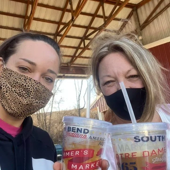Two women wearing face masks and holding large clear cups with straws. The cups display a logo with the words 'Bend Indiana America' and 'Farmer's Market'. The women appear to be standing outside on a porch with a wooden roof.