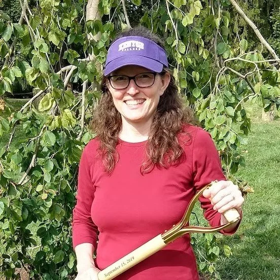 Woman in red shirt and purple cap holding garden hand tool outdoors with green leafy background.