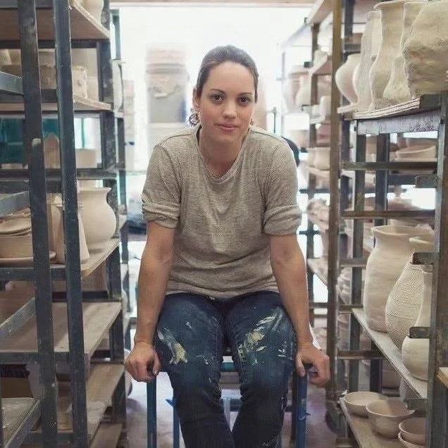 A woman sitting in a chair between shelves of pottery in a ceramics studio or store, wearing a gray shirt and paint-stained jeans.