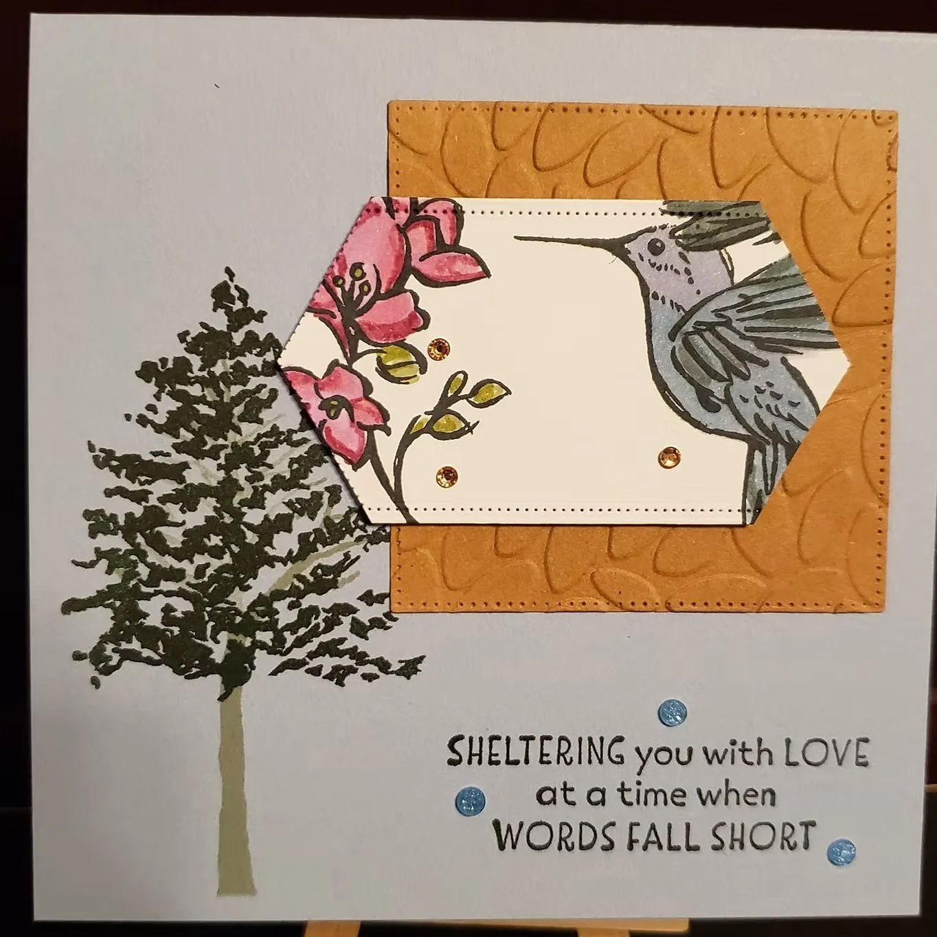 A greeting card with a tree on the left and a framed illustration of a hummingbird and pink flowers. The frame has an orange border with leaf patterns and is decorated with small shiny embellishments. Below, part of the message says, "Sheltering you with love at a time when words fall short."