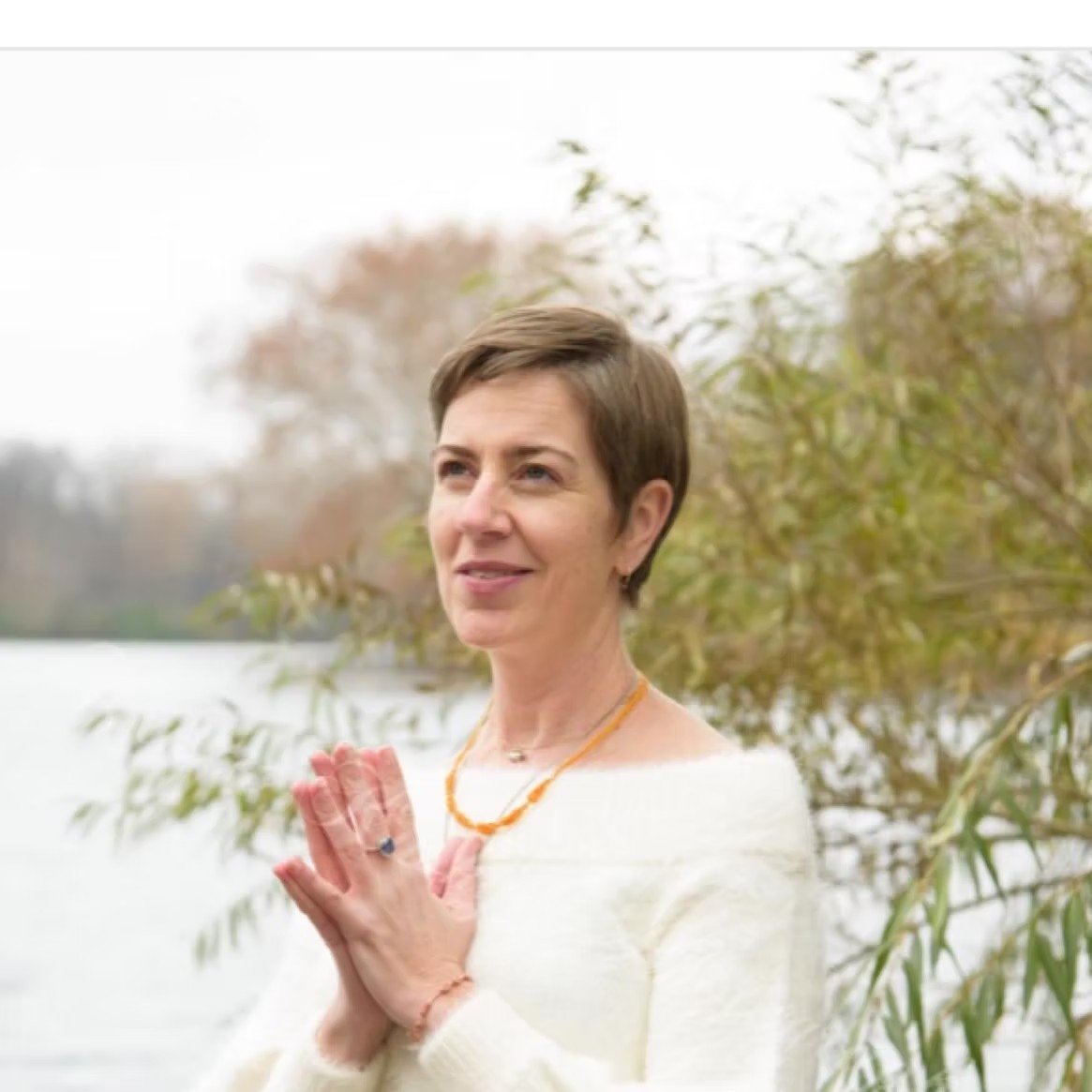 A woman with short brown hair, wearing a white sweater and an orange necklace, stands outdoors near water and trees with autumn leaves, holding her hands together in a prayer or meditative pose.