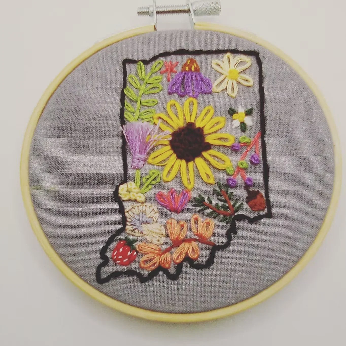 Embroidery of the outline of the state of Oklahoma with colorful floral and nature motifs, on gray fabric inside a yellow hoop.