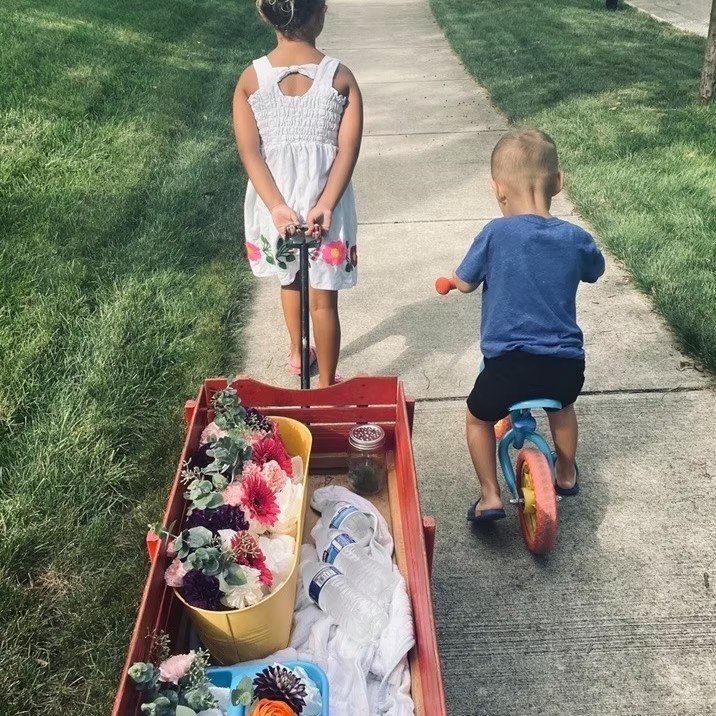 Two young children, a girl in a white dress and a baby boy in a blue shirt, walk on a sidewalk with a red wagon filled with flowers, a jar, and a towel.