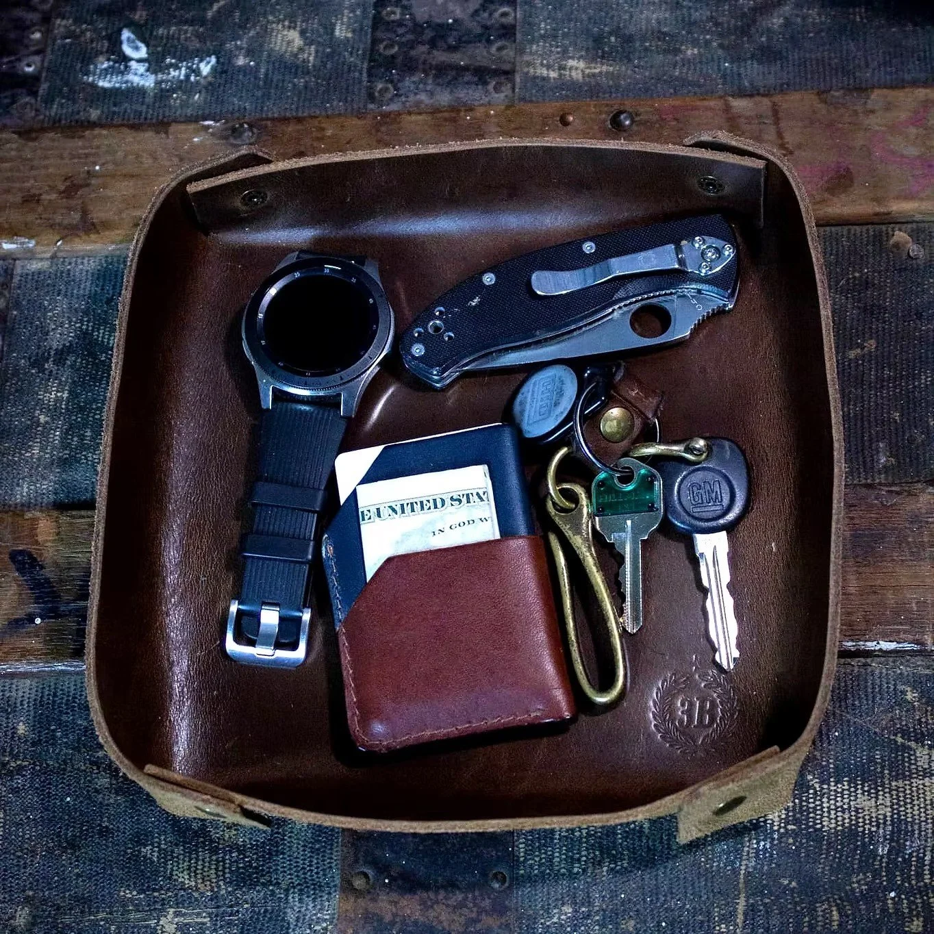 A brown leather tray containing a smartwatch, a folding pocket knife, a pair of keys with a GM key fob, a small leather wallet, a keychain with a brass hook, and a small cylindrical item, all placed on a wooden surface.