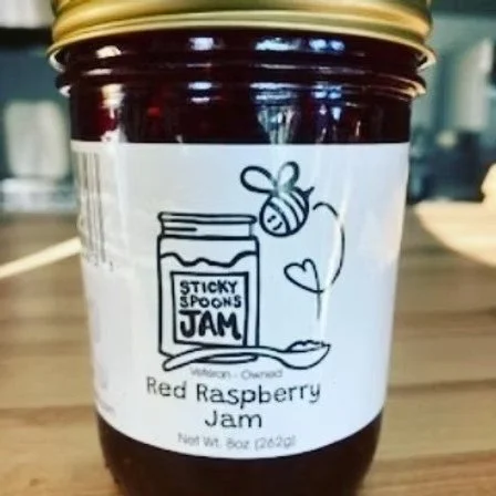 Jar of sticky spoon jam with red raspberry flavor, gold lid, and white label showing a spoon, a honey bee, and a heart.