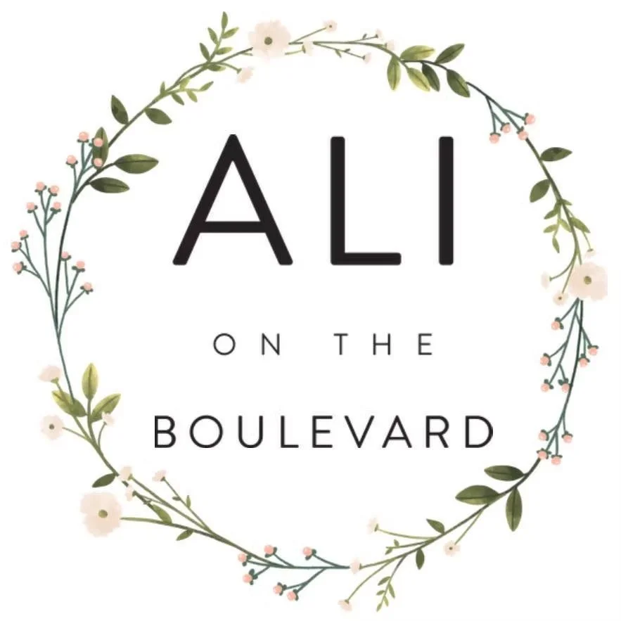Floral wreath with pink and white flowers and green leaves surrounding the text "Ali on the Boulevard" in black font.