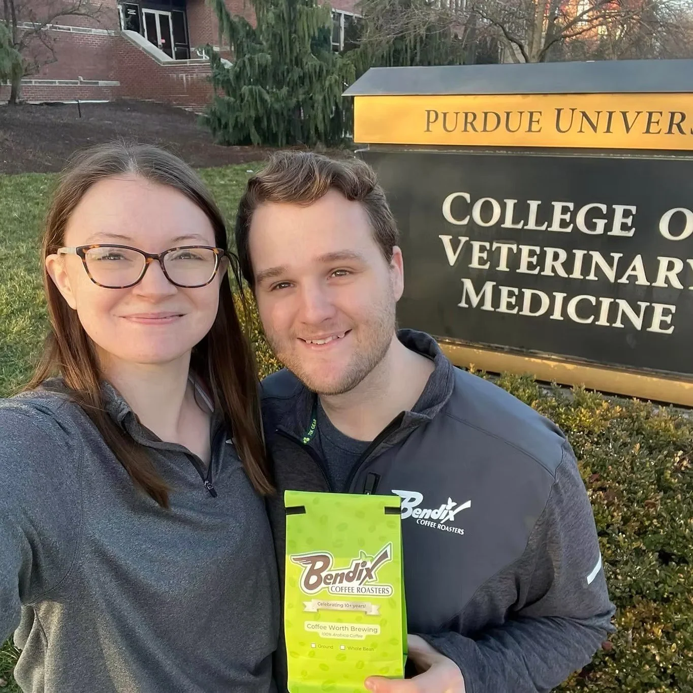 Two people standing outdoors near a sign that reads 'Purdue University College of Veterinary Medicine.' The woman on the left has long brown hair, glasses, and is smiling. The man on the right has short brown hair and a beard, and is smiling while holding a bright green packet of coffee beans from Bendix Coffee Roasters. The background includes trees and a building.