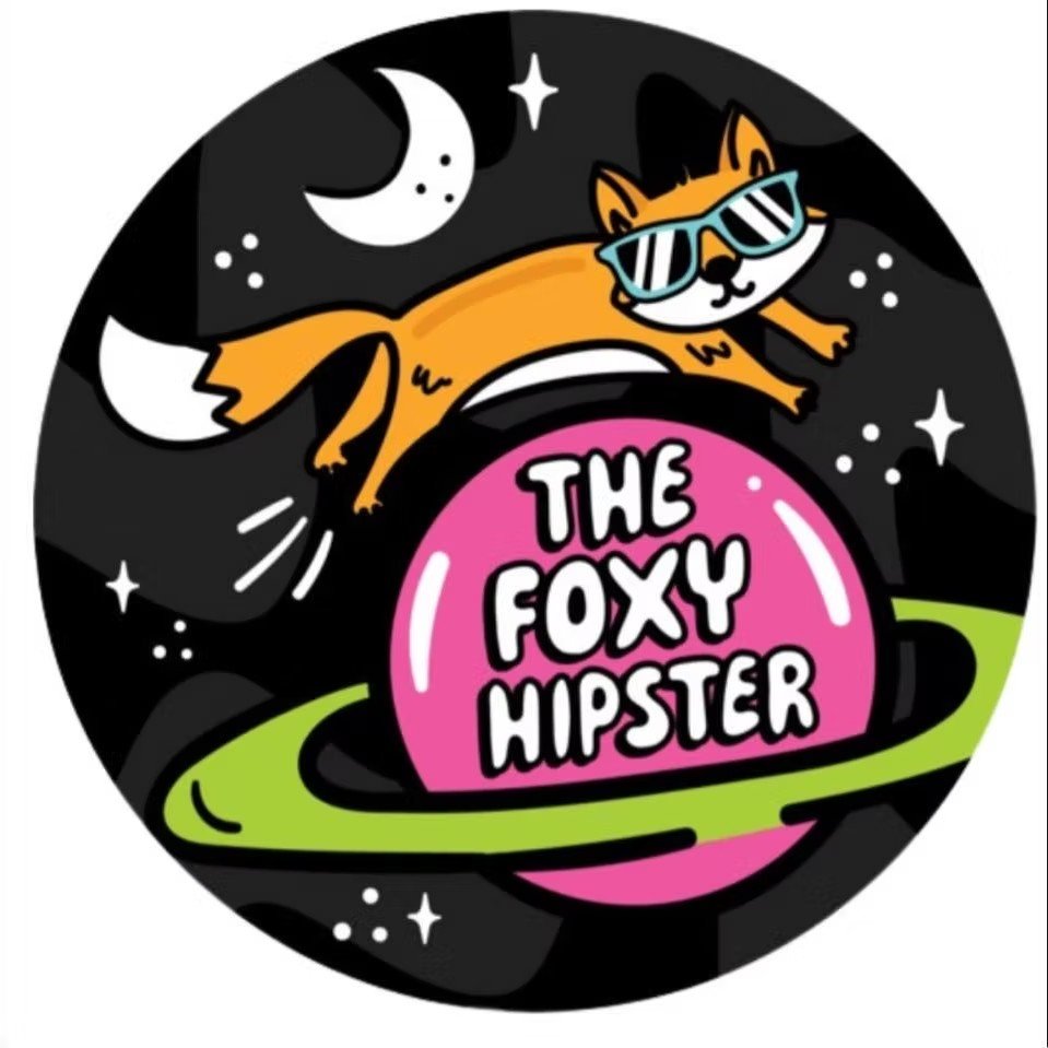 Cartoon illustration of a fox with sunglasses flying in space above a pink planet that has the text "The Foxy Hipster" on it, with stars and moons in the background.