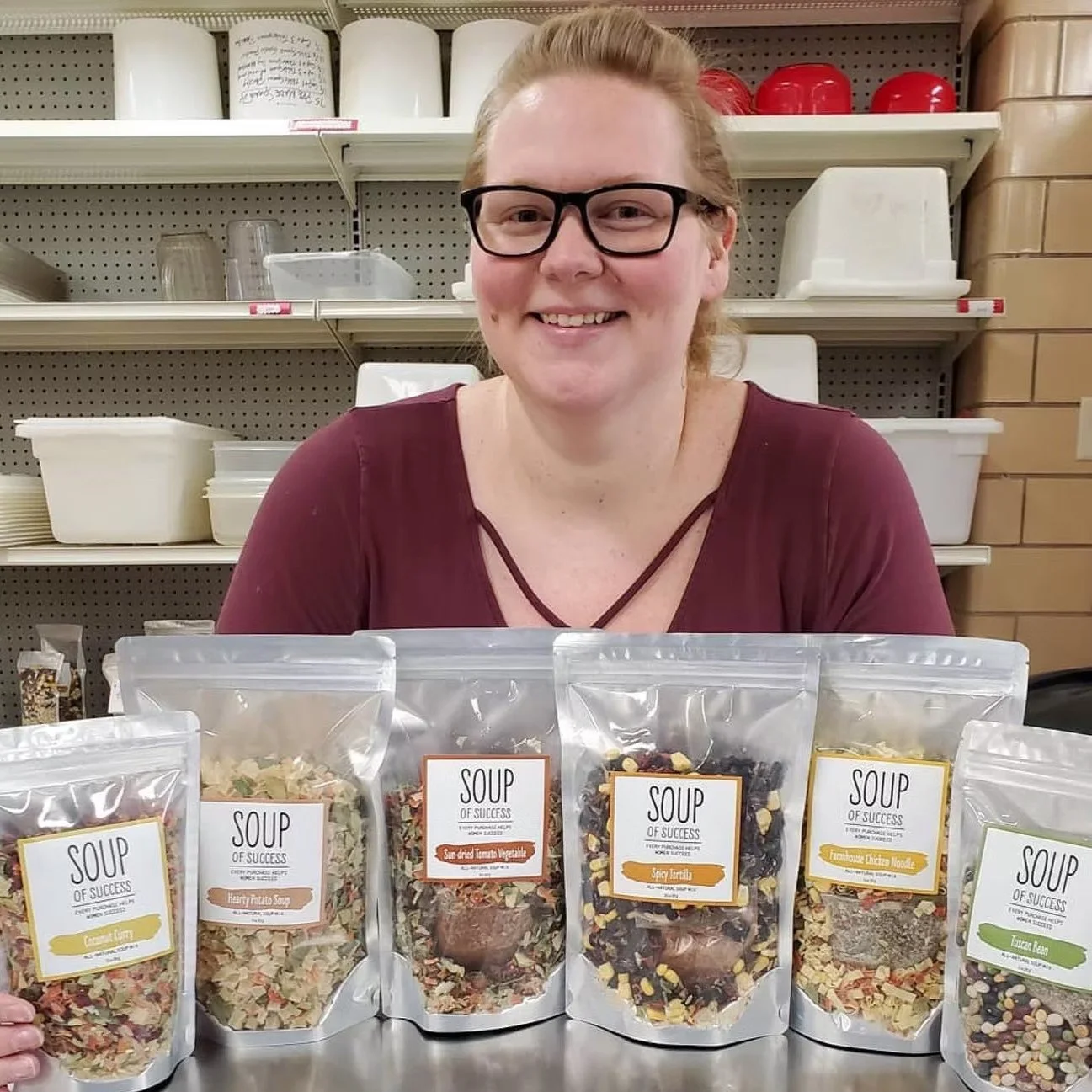 A woman with glasses smiling behind a table of packaged soup mixes labeled 'Soup of Success' in front of shelves with various containers and kitchen items.