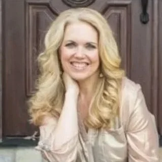 A woman with long blonde hair smiling and resting her chin on her hand in front of a wooden door.