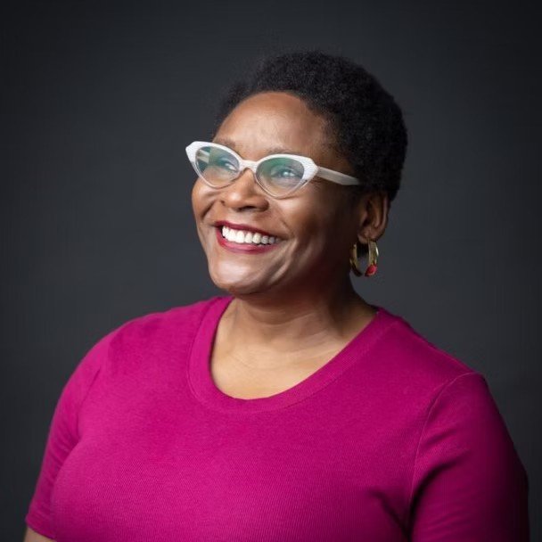 Happy woman with short curly hair wearing white glasses, gold hoop earrings, and a bright pink shirt against a dark background.
