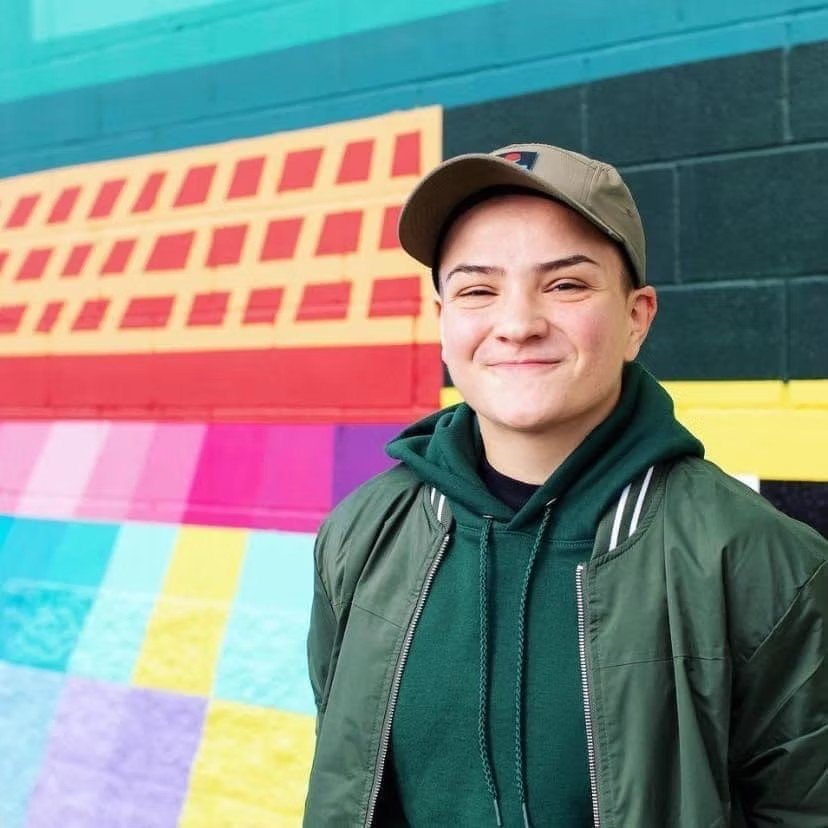 Young person smiling in front of a colorful mural that features geometric shapes and bold colors, including red, pink, yellow, and turquoise.