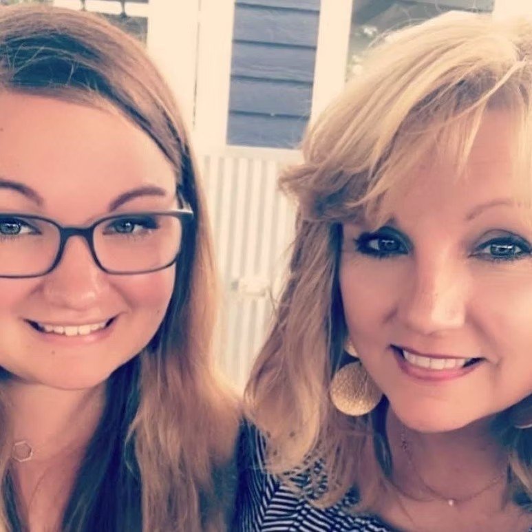 Two women smiling and taking a selfie outdoors with a building and a window in the background.