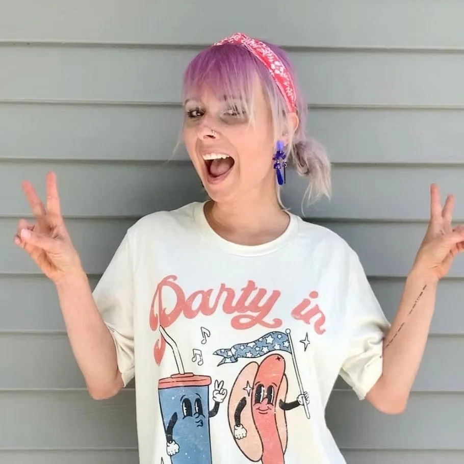 A woman with pink hair, wearing a white graphic T-shirt, pink bandana, and glowing earrings, is smiling and making peace signs with both hands while standing in front of a gray horizontal siding wall.