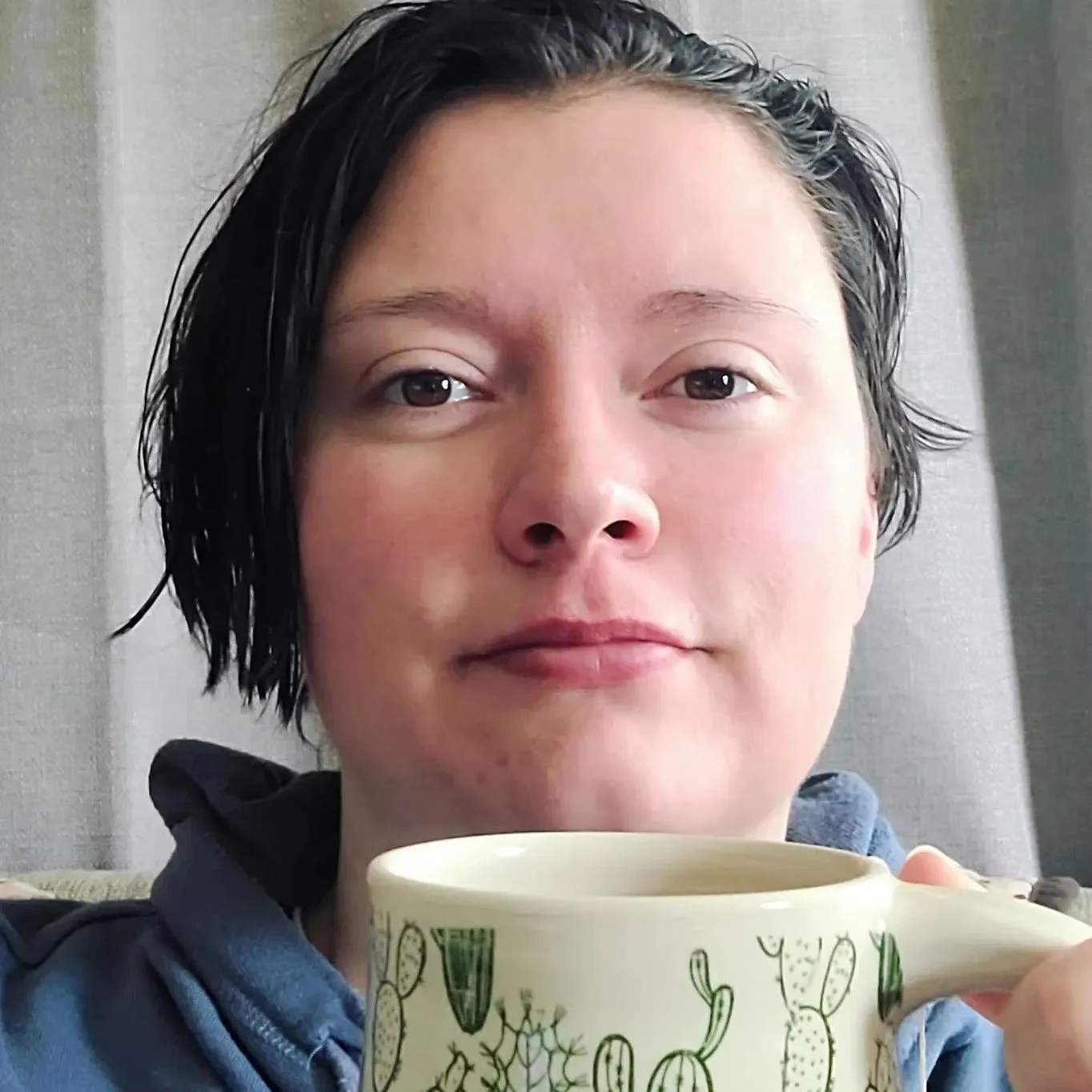 A person with short black hair holding a cup with cactus designs on it, looking at the camera.