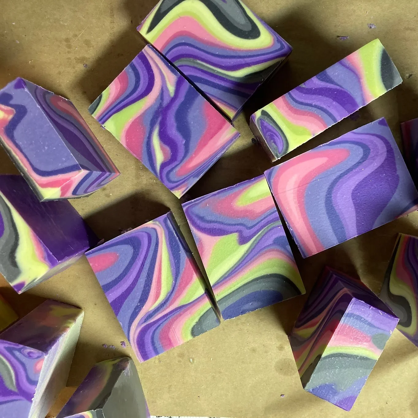 Multiple handmade soap bars with a marbled swirl pattern in shades of purple, pink, yellow, black, and gray, placed on a brown surface.