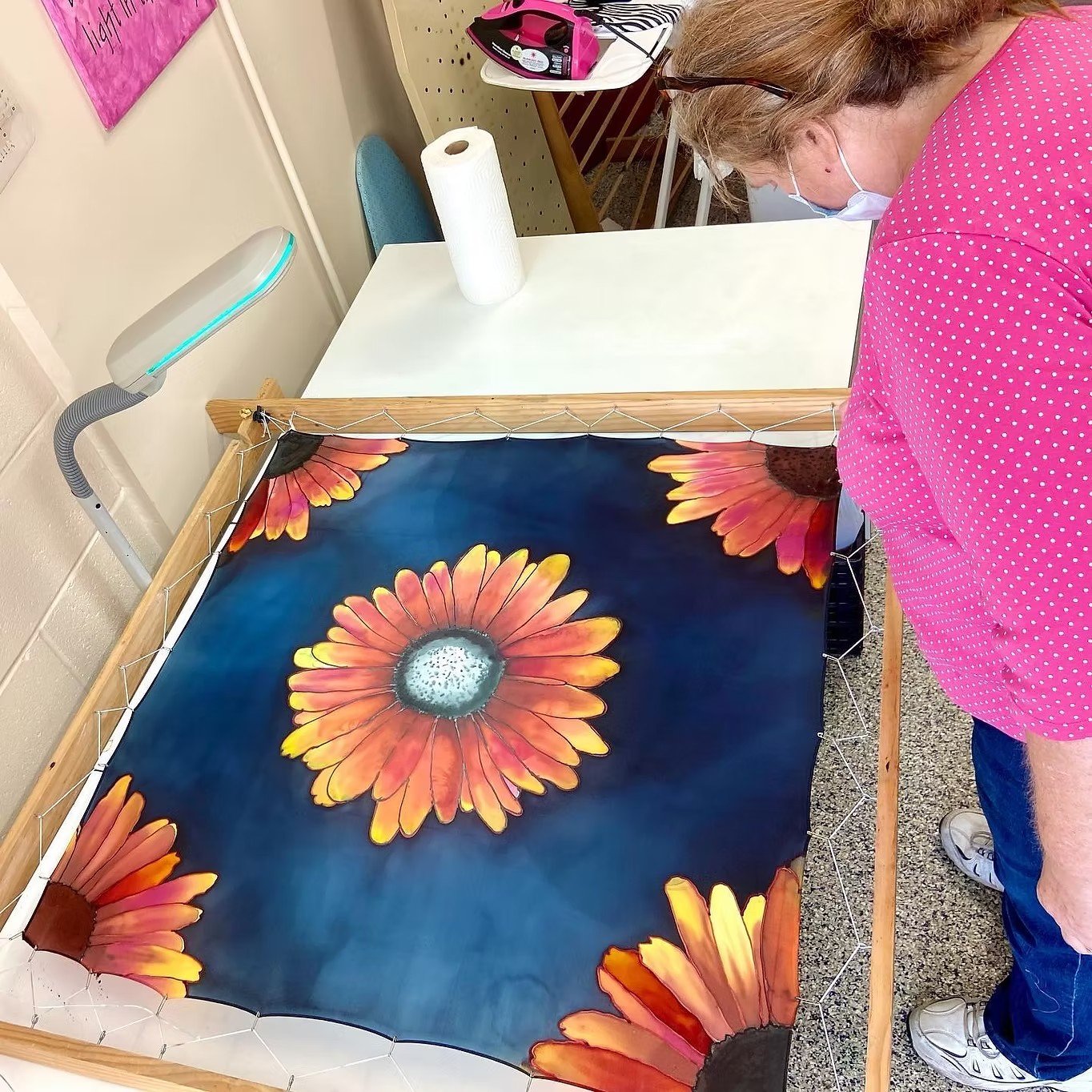 A person in a pink polka dot shirt and jeans arranging a large fabric or tapestry with orange and yellow flowers on a dark blue background in a room.