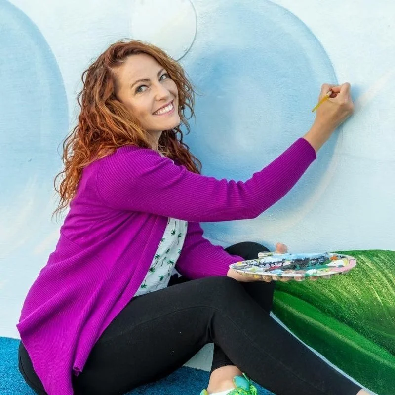 A woman with curly red hair wearing a purple cardigan and black pants, sitting on the floor and painting a large mural on a wall with blue and green colors, holding a paint palette and a brush.