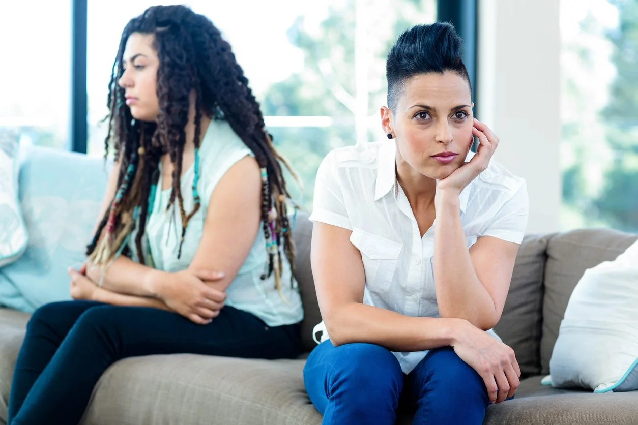 Two women sitting on a couch, appearing upset or upset with each other; one has crossed arms and looks away, the other rests her chin on her hand and looks directly at the camera.