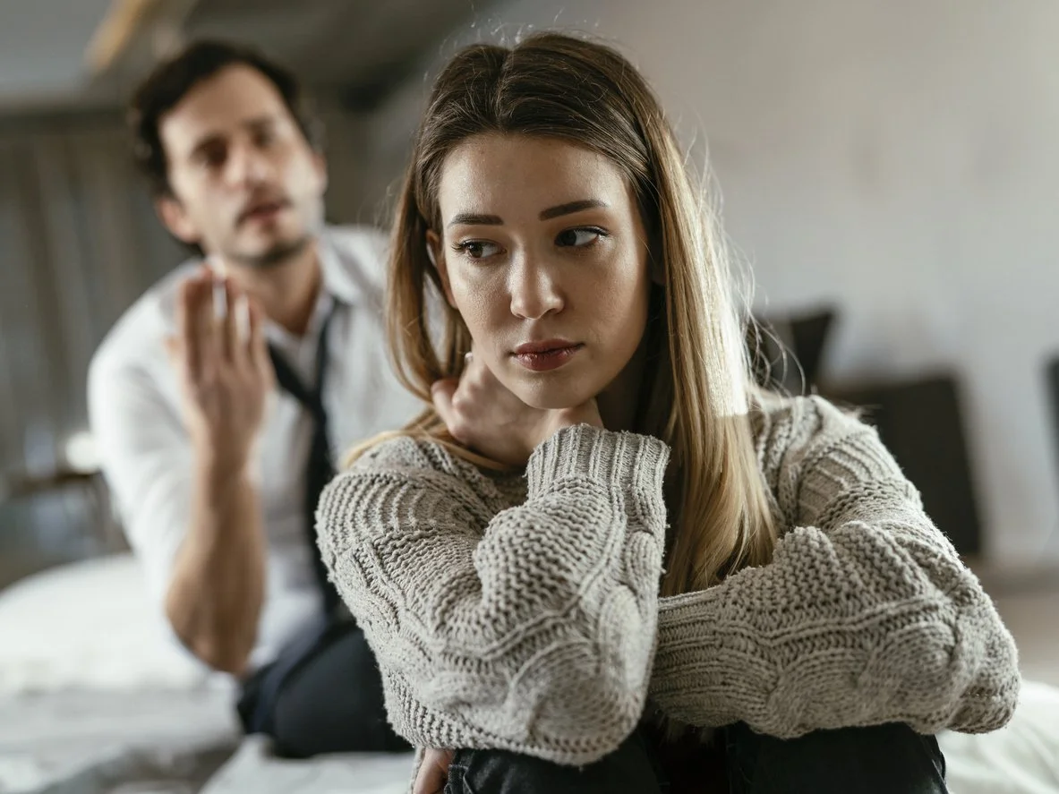 A young woman with long light brown hair sitting on a bed, looking away with a sad or thoughtful expression, wearing a beige knitted sweater. In the background, a man with dark hair and a white shirt is slightly out of focus, gesturing with his hand.