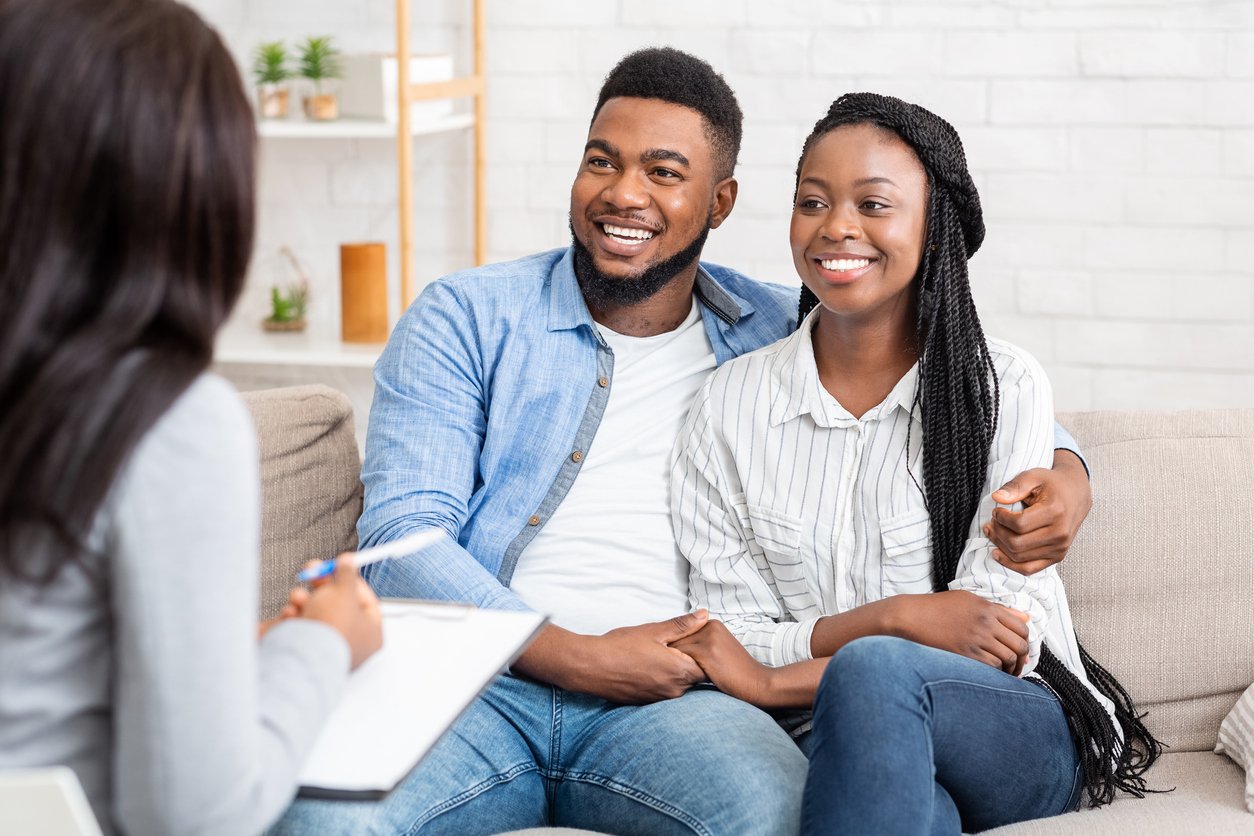 A couple sitting on a beige couch, smiling and holding hands, talking to a woman with a notepad, in a bright living room with white brick walls and decorative plants.
