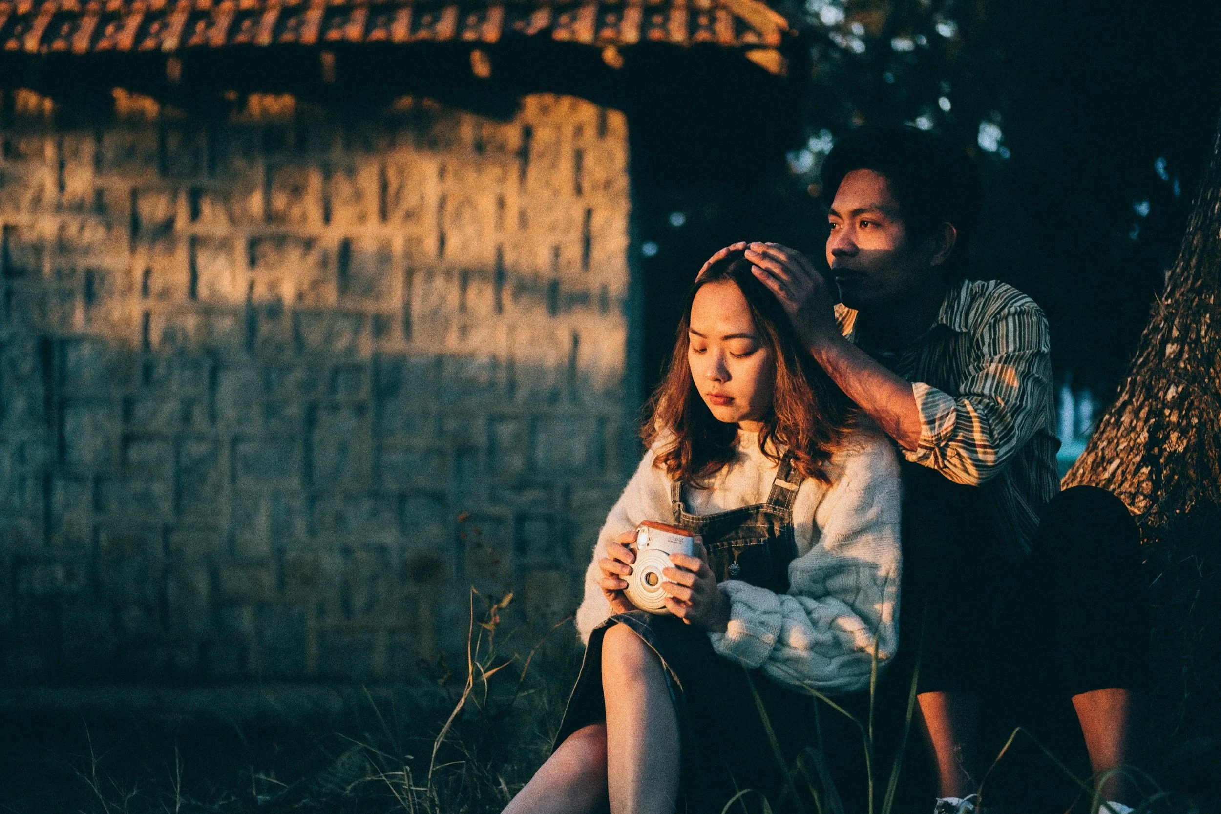 A young woman with long hair dressed in a sweater and overalls sitting outdoors at night, holding a camera. A man with dark hair in a striped shirt is behind her, gently touching her head while sitting against a tree. They are near a brick wall and surrounded by grass and trees.