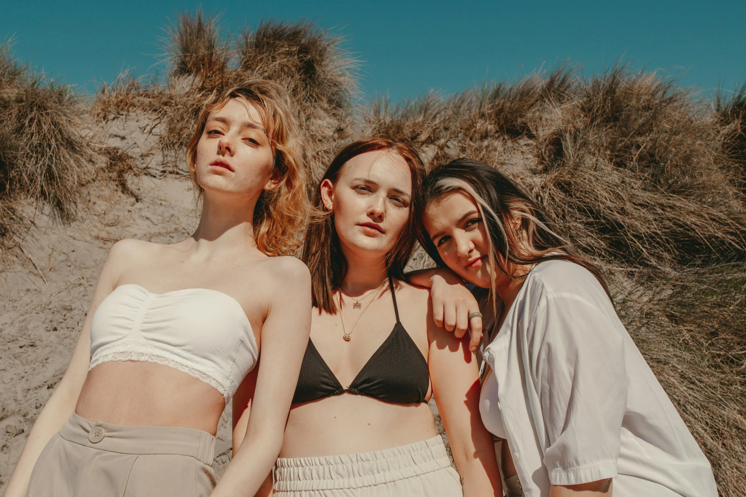 Three young women standing close together on a sandy beach with dunes and grass in the background.
