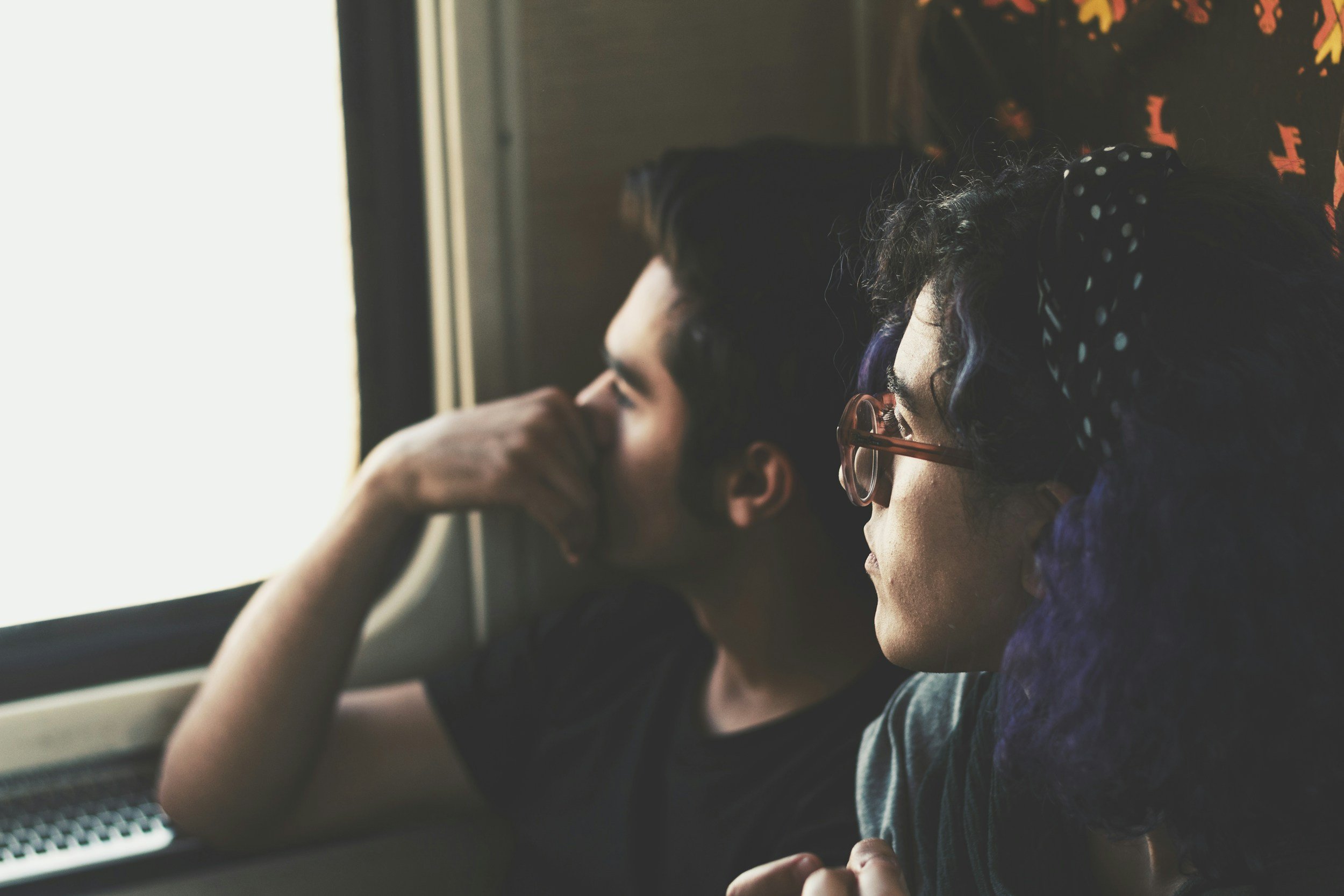 Two children, a boy and a girl, are sitting by a window on a bus or train. They are looking out the window, with the girl wearing glasses and the boy resting his head on his hand.