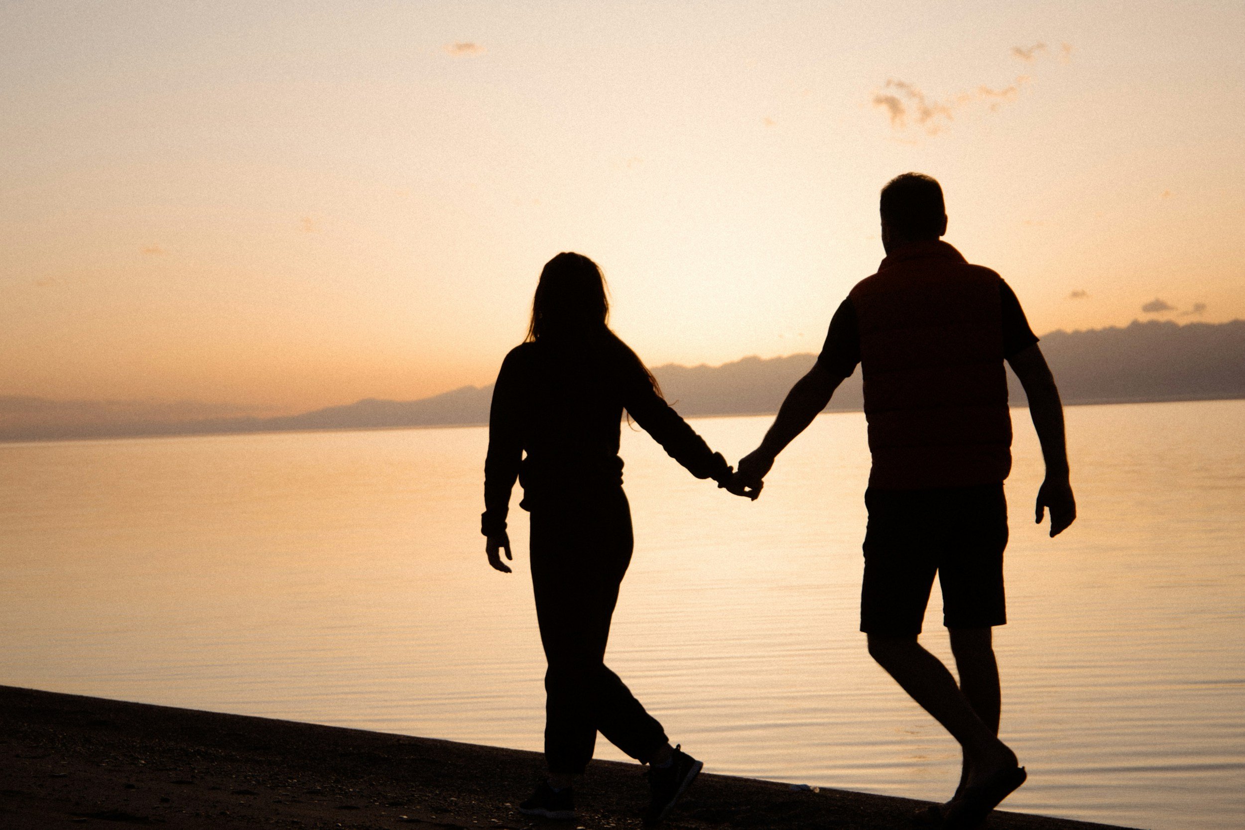 Silhouettes of a man and woman holding hands and walking along a beach at sunset.