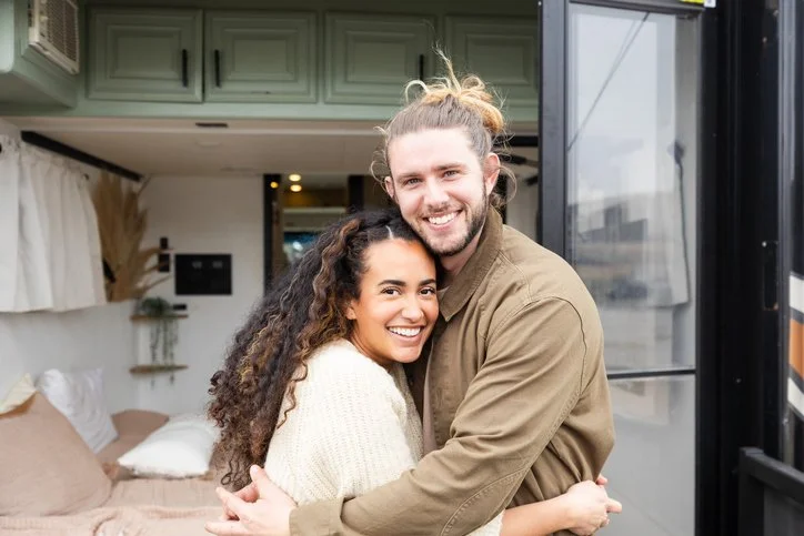 A happy couple hugging inside a tiny house or RV with a cozy bedroom in the background.