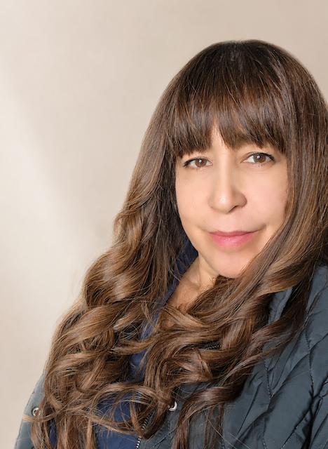 A woman with long, wavy brown hair and bangs, wearing a dark jacket, looking at the camera with a neutral expression.