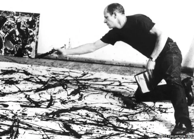 An artist in a black shirt and jeans is painting an abstract black and white artwork on a large canvas with a brush, in a studio with another painting in progress in the background.
