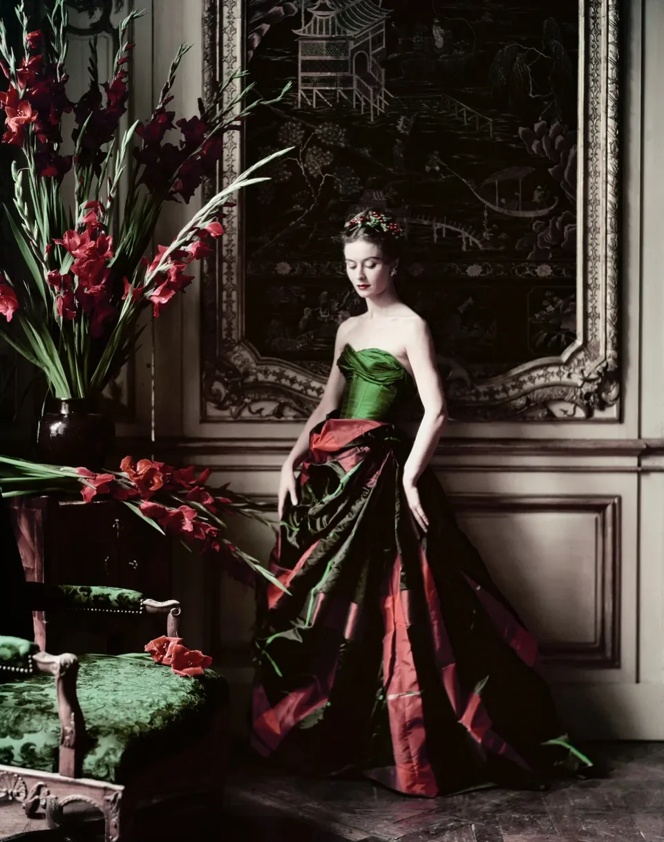 A woman wearing a green strapless bodice and a long, voluminous skirt with a black, red, and green floral pattern, standing in a dimly lit room with ornate wood paneling, a large dark tapestry on the wall, and a tall floral arrangement of red and purple flowers beside her.