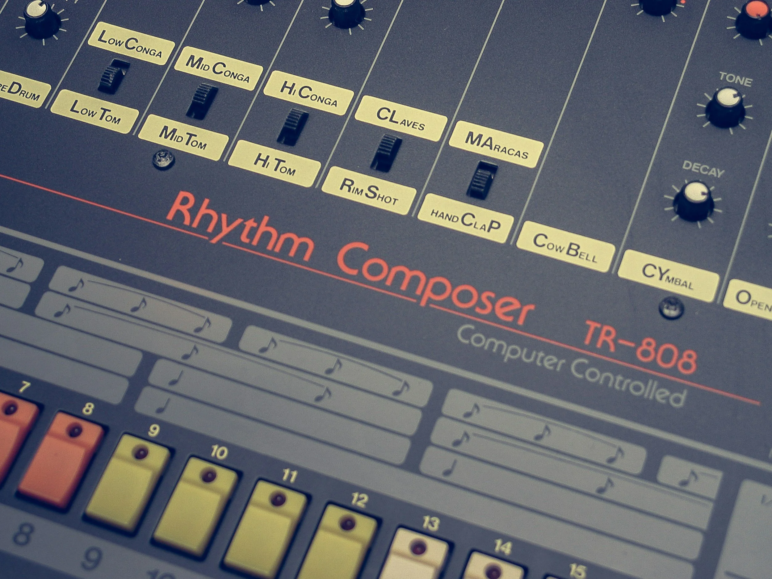 Close-up of a music production synthesizer controller with various labeled sliders and knobs, including Low Conga, Mid Conga, Hi Conga, Claves, Maracas, and others, with orange, white, and black buttons and a black background.