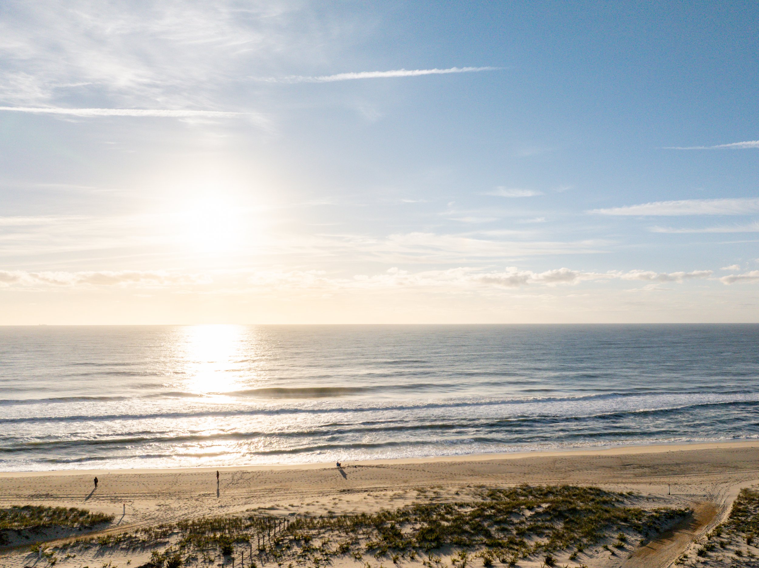Sunset over the ocean with waves crashing on the sandy beach and a few people walking along the shoreline.