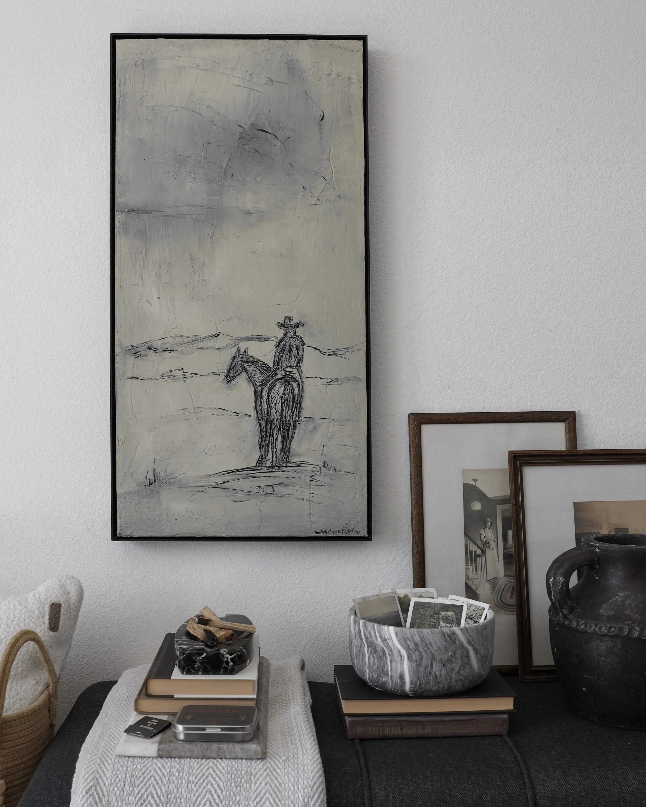 Decorative interior space with framed photographs, a large black and white painting of a cowboy on horseback on the wall, a black vase, a marble bowl filled with photographs, books, and small decor items on a table.